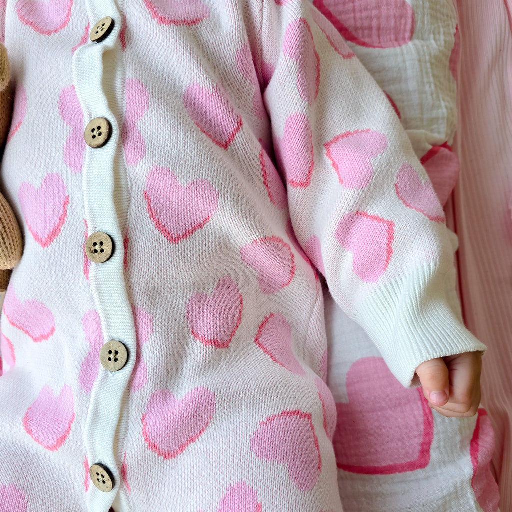 A close-up of a baby wearing a white knit one-piece romper with pink heart patterns and wooden buttons, lying on a pink and white patterned blanket.