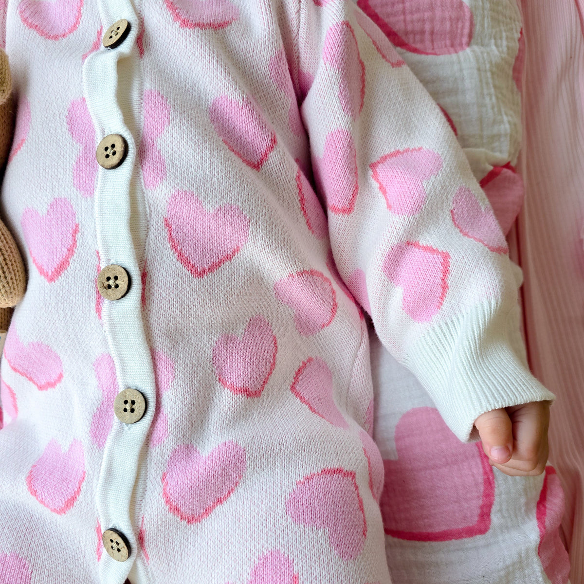 A close-up of a baby wearing a white knit one-piece romper with pink heart patterns and wooden buttons, lying on a pink and white patterned blanket.